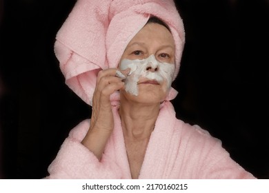 Sad Elderly Woman In Pink Bathrobe And Wrapped Towel On Head Applying Clay Mask To Face