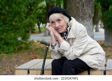 A Sad Elderly Woman With A Walking Stick Sits On A Bench Outside In Autumn. Pretty, Pensive Elderly Lady With Gray Hair On A Walk In The Park.