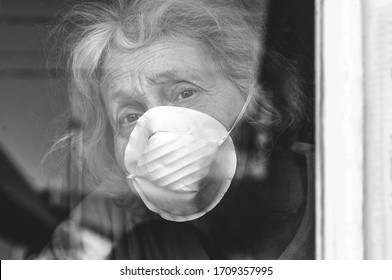 A Sad Elderly Wrinkled Woman Is Looking Through The Window In The Protective Mask Of Her House. Self-isolation Of The Elderly Coronavirus Covid-19. Quarantine And Isolation To Keep Old People Healthy