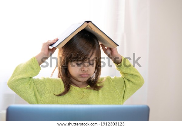 Sad elementary schooler girl studying at home\
with a book on her head because she is tired and angry - Little\
girl studying online and using internet lessons and school book -\
Digital divide concept