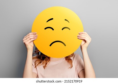 Sad emoji. Girl holds a yellow emoticon with sad face isolated on a gray background. unhappy emoji - Shutterstock ID 2152170919