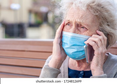 Sad Emotional Elderly Woman In Protective Mask On Her Face Plaintively Talking On Cell Phone Holding His Head In The Town. COVID-2019, Coronavirus. Senior Health. Weakening Quarantine. Outdoors