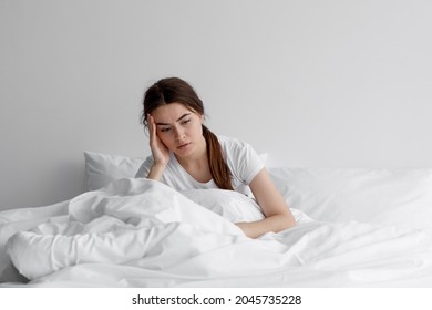 Sad european millennial female, worried, suffers from insomnia and headache. Lady sits on bed in bedroom at home, copy space. Migraines, health and mental problems, lack of sleep and depression
