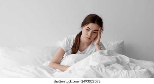 Sad european millennial female, worried, suffers from insomnia and headache. Lady sits on bed in bedroom at home, copy space. Migraines, health and mental problems, lack of sleep and depression - Powered by Shutterstock