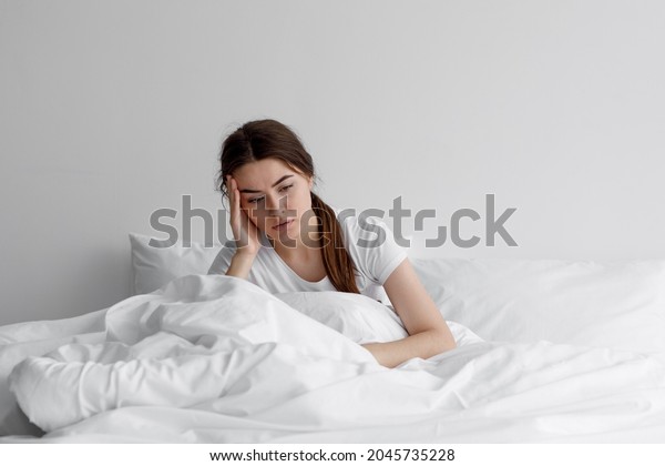 Sad european millennial female, worried,\
suffers from insomnia and headache. Lady sits on bed in bedroom at\
home, copy space. Migraines, health and mental problems, lack of\
sleep and depression