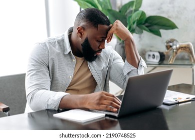 Sad Exhausted African American Young Man, Company Employee, Sitting At A Desk, Looks Tired And Stressed, Experience Headache, Migraine, Needs Rest, Closed His Eyes. Overworked From Work, Deadline