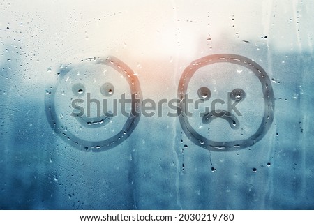 Sad face and happy smile doodle painted on window flooded with raindrops on background of city, concept of opposites