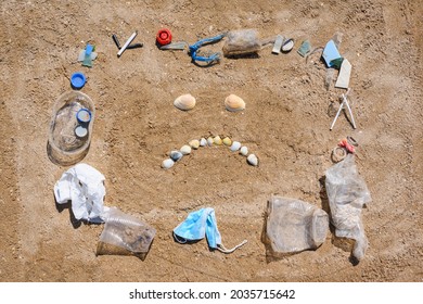 A Sad Face On The Sand Of Small Seashells Inside A Pile Of Rubbish. The Concept Of Pollution Of Nature With Household Waste.