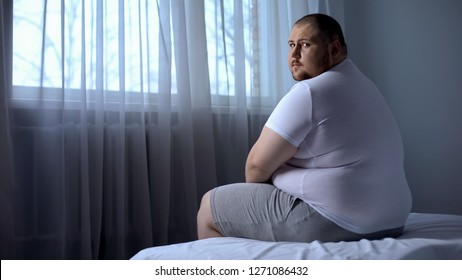 Sad fat man sitting on bed at home, looking at camera, depression, insecurities
