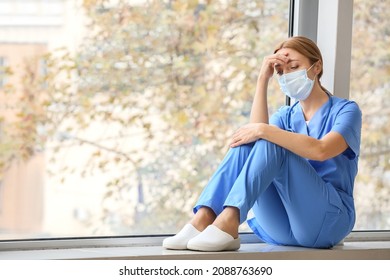 Sad Female Doctor Sitting On Window Sill In Clinic