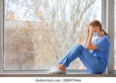 Sad Female Doctor Sitting On Window Sill In Clinic