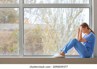 Sad Female Doctor Sitting On Window Sill In Clinic