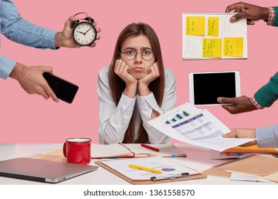Sad female workaholic keeps hands under chin, busy making project work, studies papers, wears elegant white shirt, sits at desktop, unknown people stretch hands with notes, alarm clock, smartphone - Shutterstock ID 1361558570