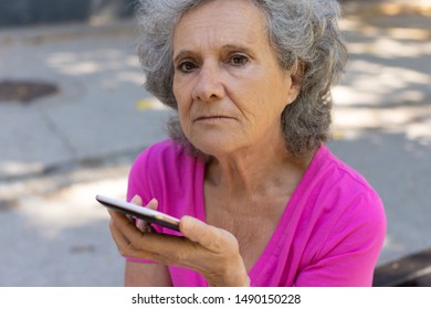 Sad Frustrated Old Lady Talking On Phone Speaker Outdoors. Upset Senior Grey Haired Woman In Casual Sitting On Park Bench And Holding Smartphone Near Face. Bad News Concept