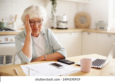 Sad frustrated senior woman pensioner having depressed look, holding hand on her face, calculating family budget, sitting at kitchen counter with laptop, papers, coffee, calculator and cell phone