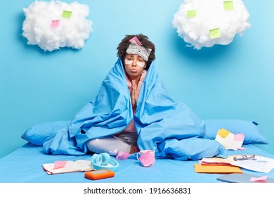 Sad Frustrated Woman With Curly Hair Keeps Palms Together Begs For Help Wrapped In Blanket Looks Unhappily Wears Blindfold On Forehead Poses On Bed Does Home Assignment Or Prepares For Test.