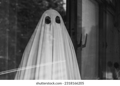 Sad ghost in white sheet outside the window of an abandoned house. Black and white photo