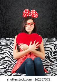 Sad Girl With A Big Hair Bow And A Heart Cushion Sitting On The Sofa And Looking At Camera