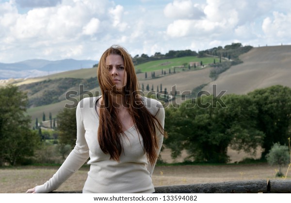 Sad girl with green eyes stands\
near fence with beautiful landscape background. Tuscan,\
Italy