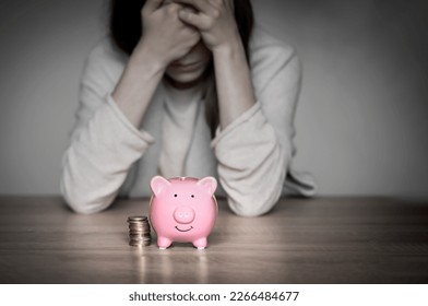 Sad girl leaned on the table, covering her face with hands, in the foreground a cute pink piggy bank. Woman lost her savings, was fired, and is experiencing financial difficulties, feels depressed.