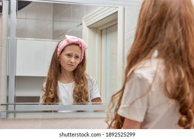 A Sad Girl Looks In The Mirror With A Frustrated Look. Long Wavy Hair. Pink Headband On Her Head.