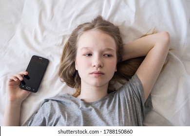 A sad girl is lying on the bed with the phone in her hands. A brooding teenager in a gray t-shirt on a white sheet. Illustration of sadness after a conversation on the phone.
