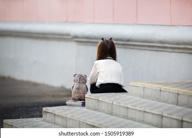 Sad girl with a plush toy sits on the steps of the school stairs with her back to the camera. Offended child. Selective focus.