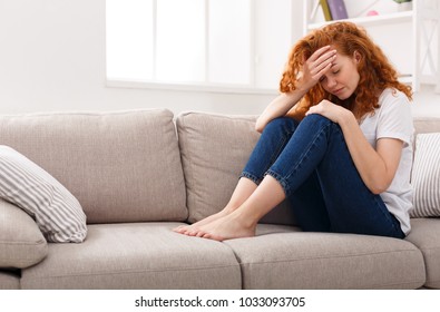 Sad girl suffering from headache. Young ginger woman feeling pain, sitting on beige sofa at home