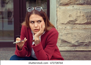 Sad girl waiting for a mobile phone call or message from her boyfriend sitting in a bench outside outdoors in the street with an urban background
