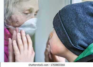 A Sad Grandmother Mature Woman In A Respiratory Mask Communicates With Grandchild Through A Window. Elderly Quarantined, Isolated. Pandemic Coronavirus Covid-19. Older People. Visit To Grandma