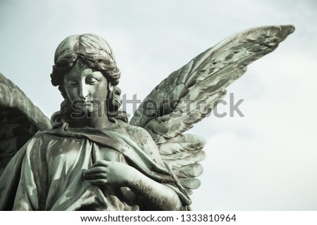 Sad guardian angel sculpture with open long wings across the frame desaturated against a bright white sky. The sad expression sculpture with eyes down and hand in front of chest. 