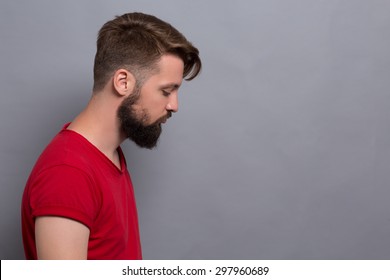 Sad Hipster Bearded Man Isolated On Grey Background. Short-haired Man In Red T-shirt Made In Profile Looking Somewhere Below.