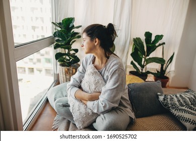 Sad hipster girl holding pillow, sitting at home during quarantine and looking at window. Stay home stay safe. Isolation at home to prevent virus epidemic. Young woman in modern room with plants