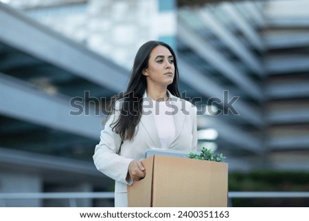 Sad Hispanic business lady carrying cardboard box with her belongings, leaving office building after layoff, reflecting economic challenges of corporate dismissal and unemployment