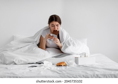 Sad hungry caucasian young lady eating chocolate bar on bed with fast food at home, suffering from stress, tension. Overeating, junk meal, depression and eating disorders. Health and mental problems