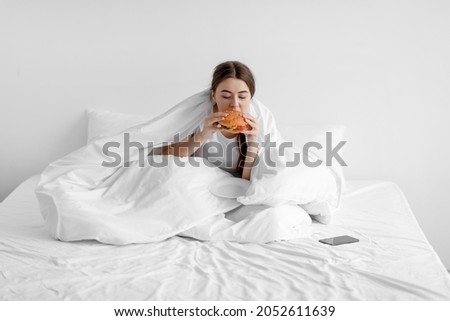 Sad hungry caucasian young woman sits on white bed at home and eating burger, suffering from depression and mental problems, empty space. Night hunger, junk food, gluttony, substitution and stress