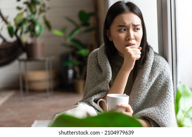 Sad ill asian girl staying on self quarantine during covid, catching flu and sitting at home with cup of tea near window, coughing in hand - Shutterstock ID 2104979639