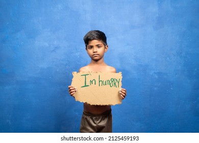 Sad Indian Poor Kid Holding I Am Hungry Sign Board By Looking At Camera On Blu Background With Copy Space - Concept Of Poverty And Malnutrition.