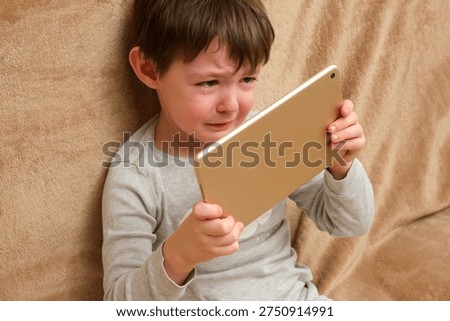 Sad kid crying while holding digital tablet on sofa. Child addiction to gadgets and screen time. Emotional developmental issues for parenting.
