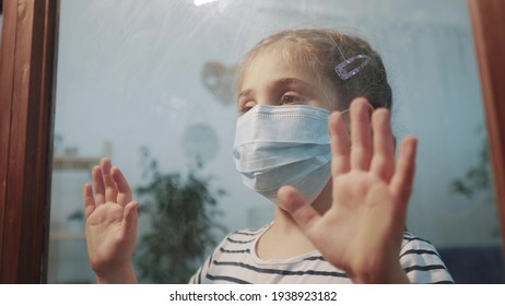 Sad Kid Girl In Protective Medical Mask By The Window Looks Out. Stay Home Coronavirus Covid 19 Concept. Sad Child In Protective Mask In Hospital Pandemic Looking Out Window. Stay Home Coronavirus