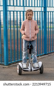 Sad Kid Girl Stand And Ride Near Spoarts Ground On Modern Eco-friendly Electric Self-balancing Scooter. Electric Hoverboard On City Street.