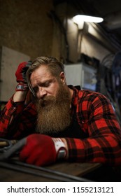 Sad Knackered Long-bearded Manual Worker In Checkered Shirt And Gloves Sitting At Table And Leaning On Hand In Workshop