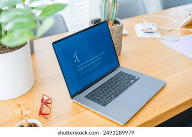 Sad laptop with blue screen error on wooden desk with plants and red glasses in the home office