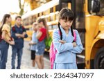 Sad little asian schoolgirl standing alone near school bus while her classmates chatting on background, upset preteen female child suffering social exclusion problem, feeling lonely and depressed