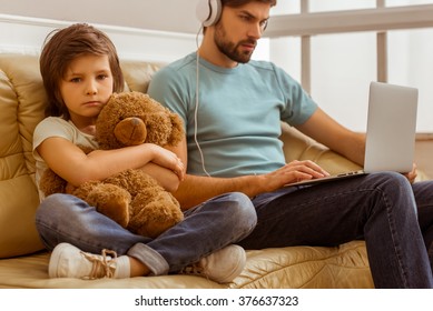 A sad little boy looking in camera and hugging a teddy bear while his father using a laptop