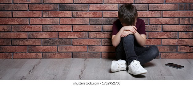 Sad Little Boy With Mobile Phone Sitting On Floor Near Brick Wall, Banner Design And Space For Text. Time To Visit Child Psychologist