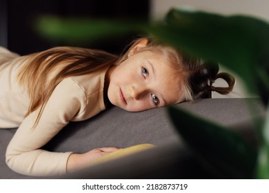 Sad Little Caucasian Girl Lying Alone On Couch In Modern Apartment And Feeling Loneliness. Bored Kid At Home During Coronavirus Covid-19 Pandemic Quarantine And Self Isolation