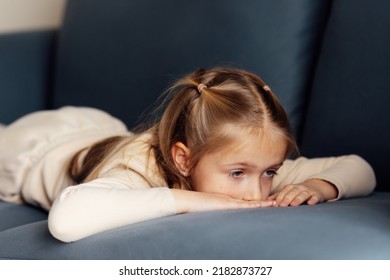 Sad Little Caucasian Girl Lying Alone On Couch In Modern Apartment And Feeling Loneliness. Bored Kid At Home During Coronavirus Covid-19 Pandemic Quarantine And Self Isolation