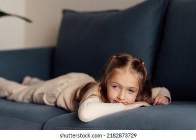 Sad Little Caucasian Girl Lying Alone On Couch In Modern Apartment And Feeling Loneliness. Bored Kid At Home During Coronavirus Covid-19 Pandemic Quarantine And Self Isolation