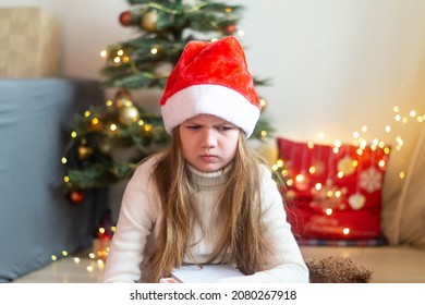 Sad Little Girl In Santa Hat Sitting On The Background Of Decorated Christmas Tree Disappointed With Gift. Cute Little Child Feeling Unhappy With Bad Holiday Present.